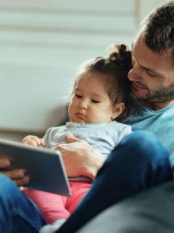 Ett barn sitter med sin pappa i en soffa och spelar på en touch-skärm.
