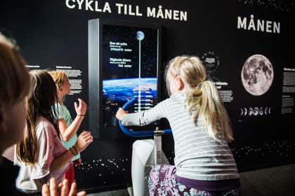 En grupp med äldre barn utforskar hur långt det skulle ta att cykla till månen vid en monter inne i utställningen Rymdresan.
